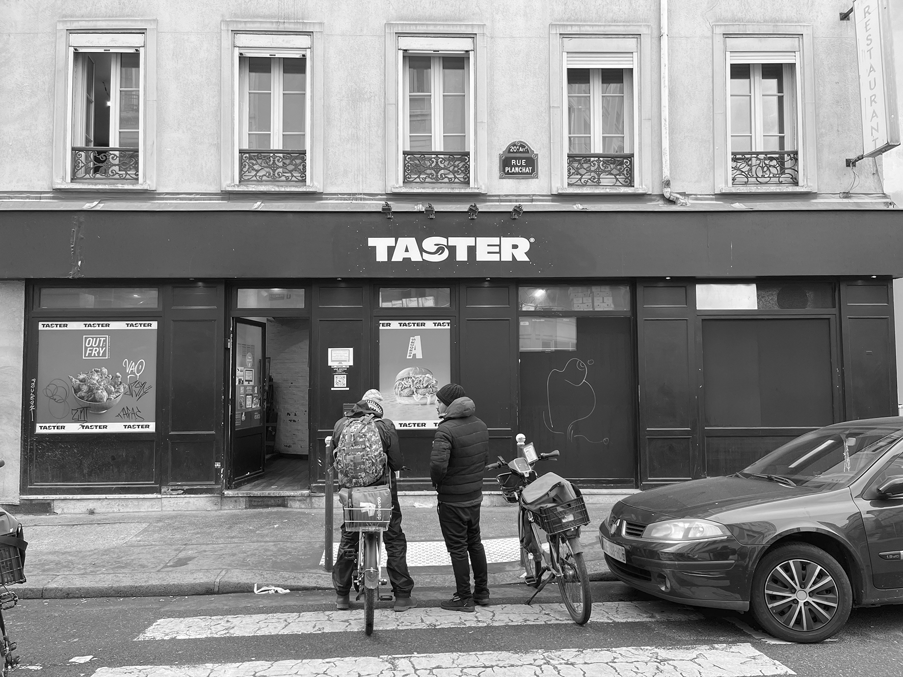 Livreurs, équipés de leurs vélos et de leurs sacs isothermes attendant leurs commandes un jeudi à 14h. La façade fonctionne comme un écran opaque : les affiches et le QR code renvoient visuellement au site internet Taster : une offre de plats cuisinés à commander en ligne et à se faire livrer. Depuis la rue, l’activité interieure est invisibilisée. Cette photographie saisit précisément l’articulation entre l’invisibilité de la production alimentaire et la visibilité contrainte des corps qui en assurent la distribution. Ces livreurs, leurs vélos et leurs sacs, incarnent ces corps et objets ressources mobilisés par l’économie des dark kitchens. Le contraste entre le bâti haussmannien et cette façade standardisée met en lumière les ruptures spatiales et sociales générées par ces nouveaux modèles économiques dans le tissu urbain traditionnel.